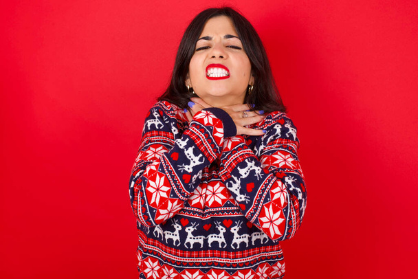 Brünette kaukasische Frau in Weihnachtspullover über rotem Hintergrund schreit ersticken, weil schmerzhafte Würgen. Gesundheitsprobleme. Asphyxid- und Selbstmordkonzept. - Foto, Bild