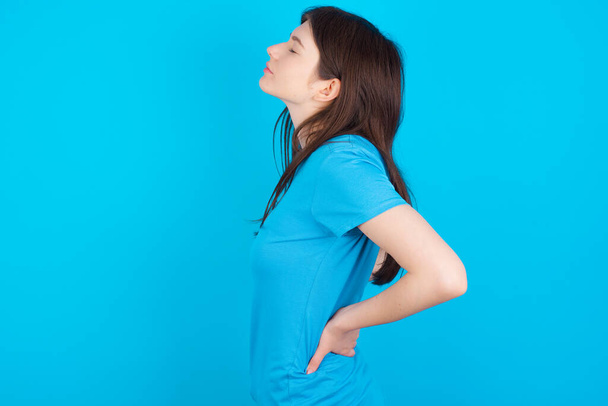 joven caucásico chica usando azul camiseta aislado sobre azul estudio fondo tiene dolor de espalda - Foto, Imagen