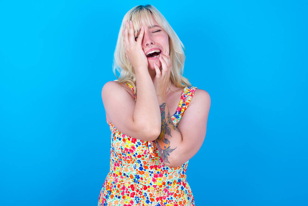 Young gloomy caucasian girl wearing floral dress isolated over blue background hiding face with hands pouting and crying, standing upset and depressed complaining about job problem. - Photo, Image