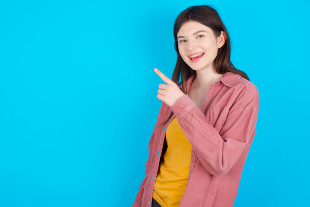молодая кавказская девушка в розовой рубашке на голубом фоне, указывая на тебя и улыбаясь. Посмотри туда.! - Фото, изображение