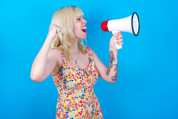 біла дівчина в квітковій сукні ізольована на синьому фоні спілкується з криком вголос, тримаючи мегафон, висловлюючи успіх і позитивну концепцію, ідею для маркетингу або продажів. - Фото, зображення
