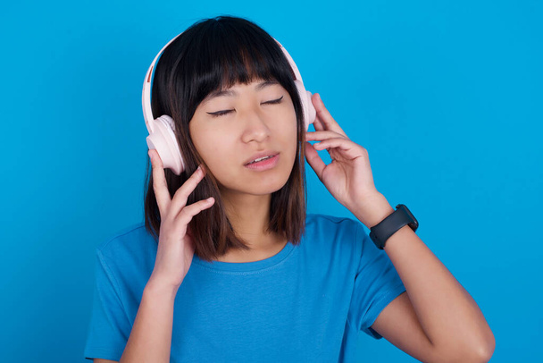 junge Asiatin trägt blaues T-Shirt vor blauem Hintergrund mit Kopfhörer auf dem Kopf, hört Musik, genießt Lieblingssong mit geschlossenen Augen, hält die Hände am Headset. - Foto, Bild