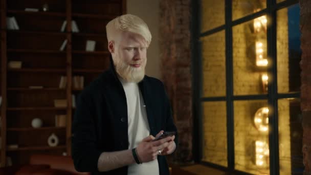 Focused albino businessman typing message on smartphone in loft office with night city lights seen in large window on motion background. Male hipster mobile phone. Casual freelancer working at night - Footage, Video