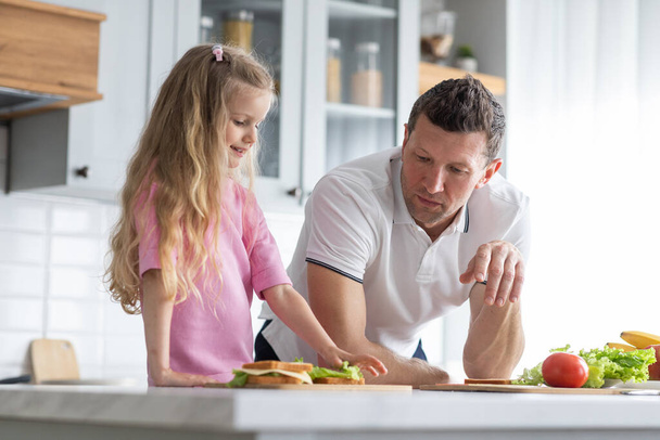 A friendly family from father with daughter stand and manage in the kitchen. A young serious man dad gives his child culinary instructions to make sandwiches independently. The concept of mutual - Photo, Image