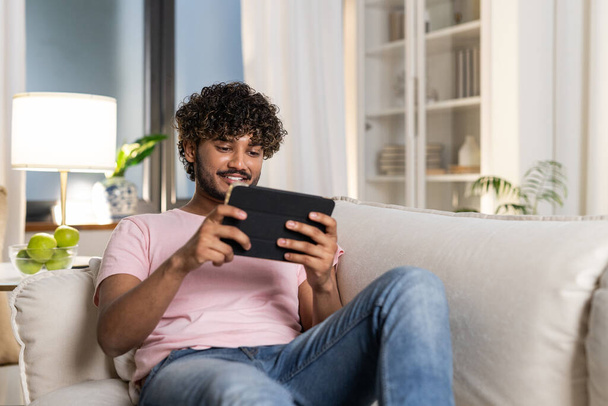 A smiling mestizo guy reclining on the sofa in the room, enjoys relax after work or study in the cozy home environment. A positive young african or american man holds a tablet in hands and watch a - Foto, Imagem