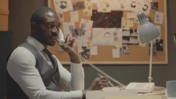 Средний снимок профессионального афроамериканского детектива-мужчины, сидящего за столом в офисе и говорящего по стационарному телефону во время расследования уголовного дела - Кадры, видео