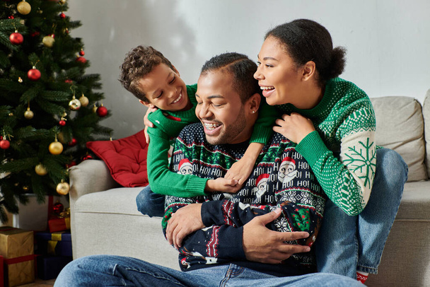 onnellinen afrikkalainen amerikkalainen perhe viihtyisissä villapaidoissa halaamassa lämpimästi ja hymyillen iloisesti, joulu - Valokuva, kuva