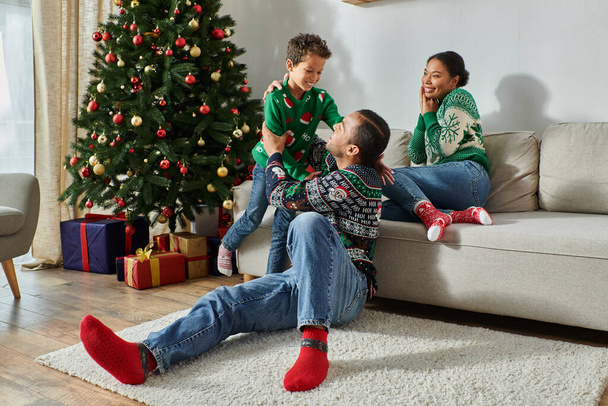 mujer afroamericana bonita mirando felizmente a su marido y a su hijo al lado del árbol de Navidad - Foto, Imagen