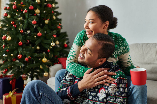 счастливая африканская американская пара обнимается тепло с чашкой кофе в руке, глядя в сторону, Рождество - Фото, изображение
