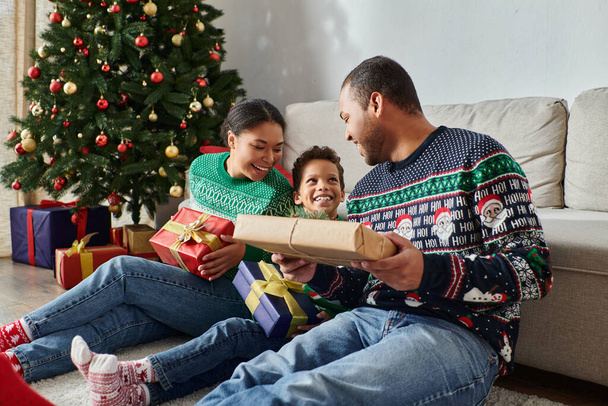 Χαρούμενη αφροαμερικάνικη οικογένεια που κάθεται στο πάτωμα κρατώντας δώρα και χαμογελώντας χαρούμενα, Χριστούγεννα - Φωτογραφία, εικόνα