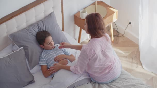Ένα άρρωστο αγόρι αισθάνεται αδιαθεσία, μετρά τη θερμοκρασία, βρίσκεται στην κρεβατοκάμαρα και διατηρεί ένα κρεβάτι ανάπαυσης. Μια στοργική νεαρή μητέρα κάθεται κοντά, δίνει σε ένα γιο ψηφιακό θερμόμετρο και αγγίζει το μέτωπό του για έλεγχο - Πλάνα, βίντεο