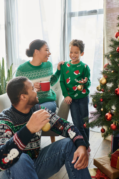 クリスマスツリーを飾る彼女の夫と息子を見てコーヒーカップを持つアフリカ系アメリカ人女性 - 写真・画像