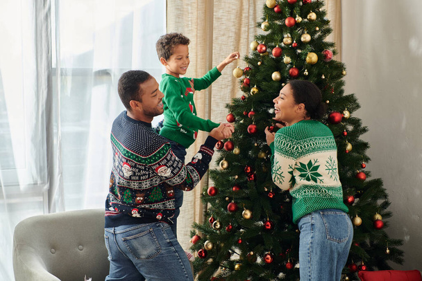 見栄えの良いアフリカ系アメリカ人家族 暖かいセーターで一緒にクリスマスツリーに問題を置く - 写真・画像