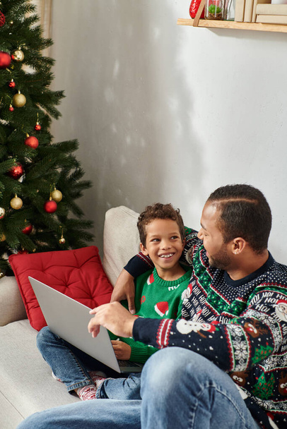 vertical shot of cheerful african american man hugging son while watching Christmas movie on laptop - Photo, Image