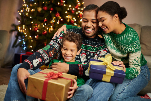 cheerful african american parents hugging their son while he holding present and smiling joyfully - Foto, Imagem
