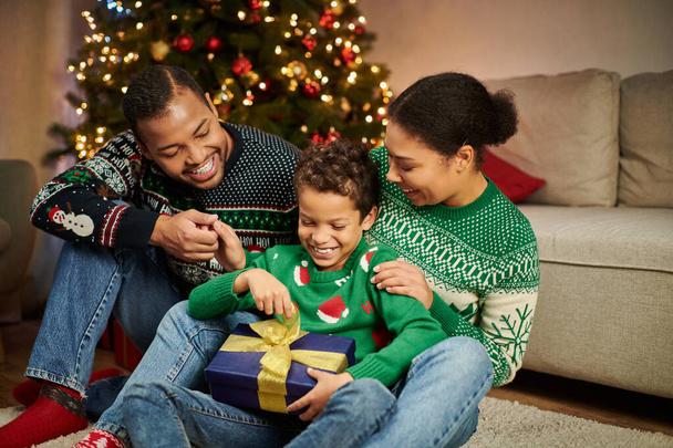 gelukkig Afrikaans amerikaanse ouders in gezellige truien kijken hoe hun zoon uit te pakken kerstcadeau - Foto, afbeelding