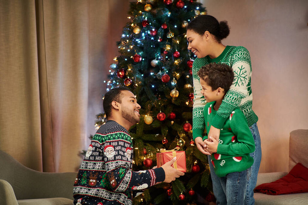bien parecido jolly africano americano hombre sosteniendo presente y mirando a su esposa e hijo, Navidad - Foto, imagen