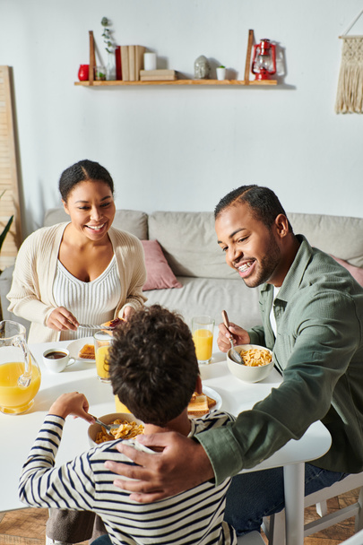 verticaal schot van gelukkig modern Afrikaans Amerikaans gezin eten ontbijt en vrolijk glimlachen - Foto, afbeelding