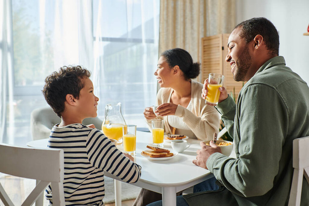 alegre adorável menino afro-americano sorrindo para seu pai, enquanto sua mãe olhando para longe, café da manhã - Foto, Imagem