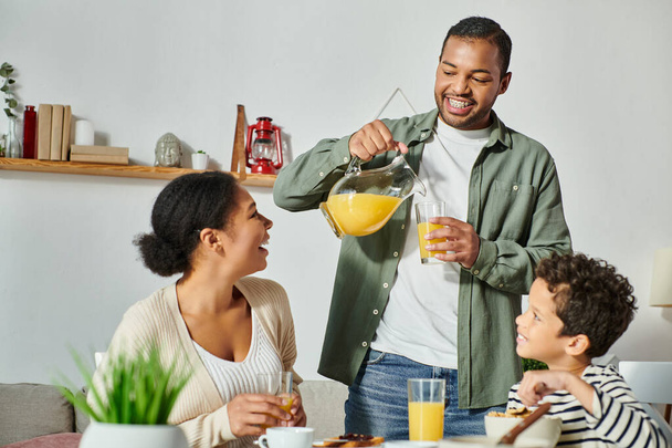 Веселый африканский американец наливает апельсиновый сок в стакан, в то время как его жена и сын улыбаются ему - Фото, изображение