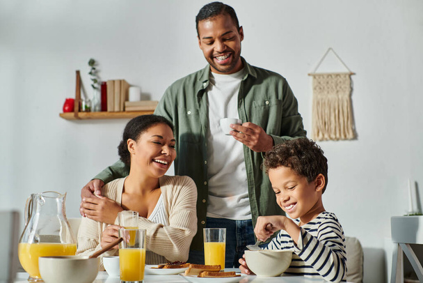 fröhliche afrikanisch-amerikanische Eltern, die ihren kleinen Sohn am Frühstückstisch liebevoll und lächelnd betrachten - Foto, Bild