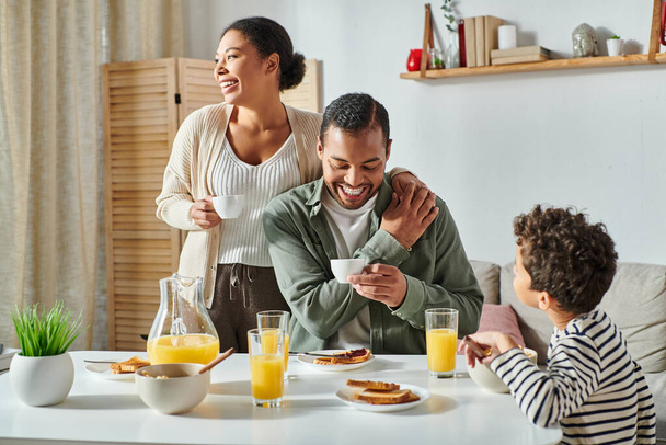 padres afroamericanos alegres con tazas de café en las manos sonriendo cerca de su hijo desayunando - Foto, imagen