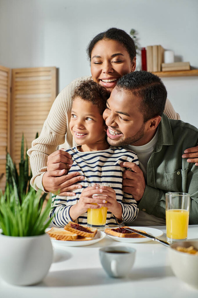 tiro vertical de família afro-americana alegre abraçando calorosamente uns aos outros na mesa de café da manhã - Foto, Imagem