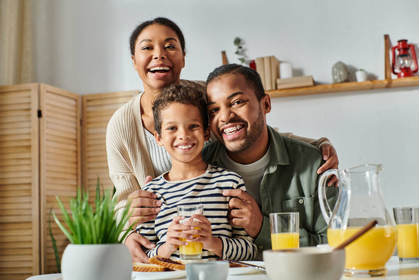 joyeuse famille afro-américaine étreignant chaleureusement et souriant joyeusement à la caméra à la table du petit déjeuner - Photo, image