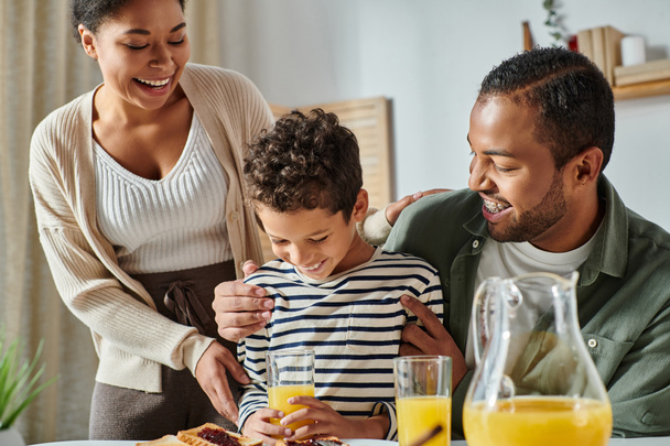 wesoła, nowoczesna afrykańska rodzina przytula się i uśmiecha przy śniadaniu, sok pomarańczowy w dłoniach - Zdjęcie, obraz