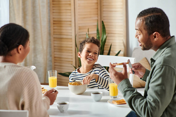 focus on happy adorable african american boy looking cheerfully at his parents at breakfast table - Photo, Image