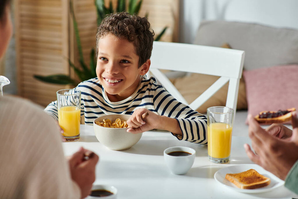 padres afroamericanos desayunando juntos con su alegre hijito comiendo copos de maíz - Foto, Imagen