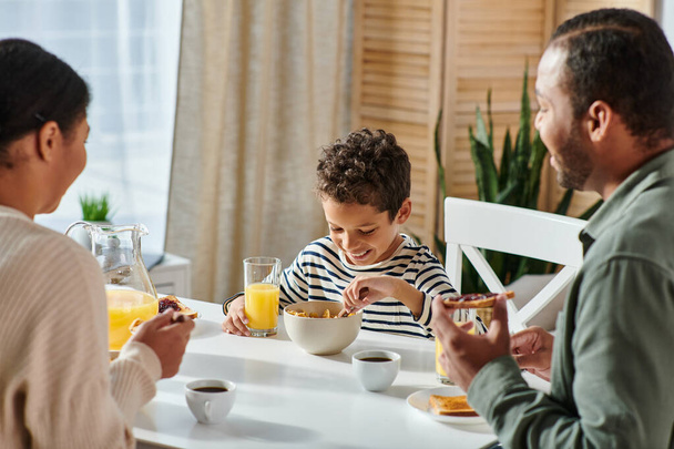 focus op gelukkig Afrikaans amerikaans jongen genieten van ontbijt en zijn wazig ouders kijken naar hem - Foto, afbeelding