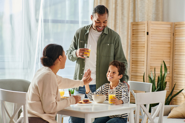 belle famille afro-américaine moderne donnant haute cinq à la table du petit déjeuner, jus d'orange à la main - Photo, image