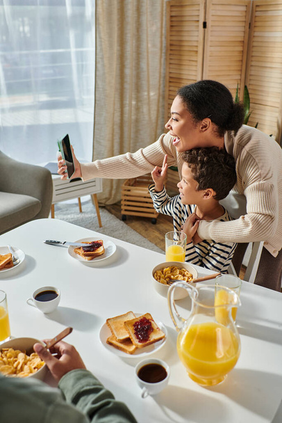 vertical shot of cheerful african american mother taking selfie at breakfast table with her son - Photo, Image