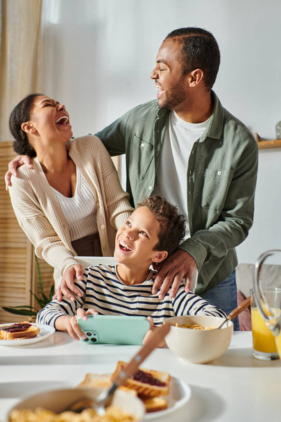 pystysuora laukaus kaunis afrikkalainen amerikkalainen perhe nauraa vilpittömästi aamiaisella, puhelin käsissä - Valokuva, kuva