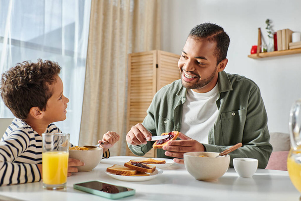 Hyvännäköinen afrikkalainen amerikkalainen isä levittää hilloa paahtoleivälle ja hymyilee pojalleen aamiaisella - Valokuva, kuva
