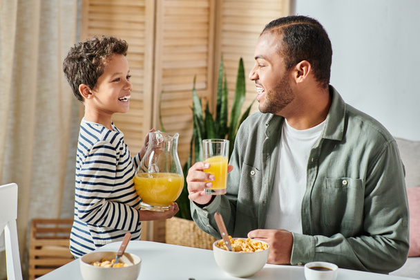 alegre afroamericano chico con jarra de jugo de naranja sonriendo a su padre en mesa de desayuno - Foto, imagen
