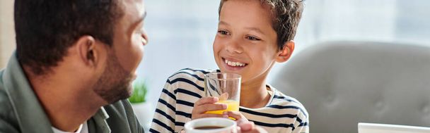 alegre lindo africano americano chico con jugo de naranja sonriendo a su padre con café, pancarta - Foto, imagen