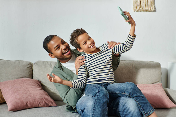 joyous african american father and son in homewear having fun and taking selfies sitting on sofa - Фото, зображення
