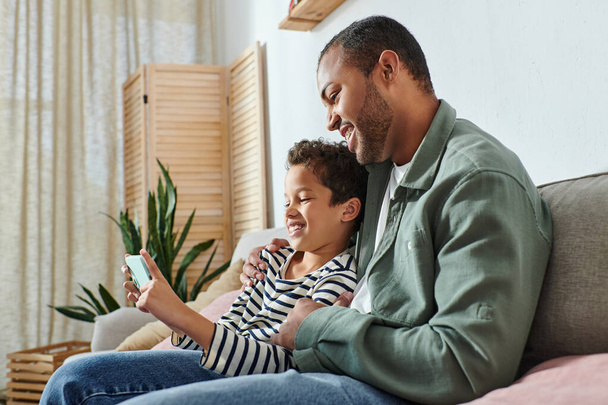 feliz padre afroamericano sentado en el sofá con su hijo abrazando y sonriendo en el teléfono móvil - Foto, Imagen