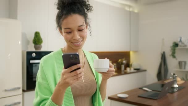 Diverse model curly woman drinking coffee and holding smartphone making food delivery order on-line, choose goods on internet slow mo. Beautiful young diverse model using smartphone in modern kitchen - Footage, Video