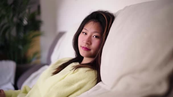 Młoda atrakcyjna Azjatka spokojne oczy relaksujący oddech siedzi na kanapie w salonie. Chińska ładna dziewczyna patrzy na beztroski aparat cieszący się wolnym czasem w domu. Kobieta przytulne życie domowe. - Materiał filmowy, wideo