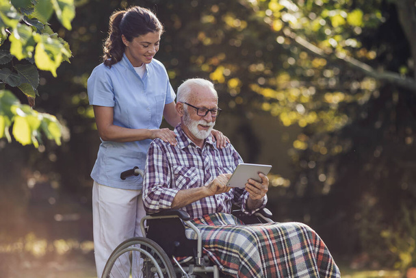 Homem sênior em cadeira de rodas mostrando algo em tablet para jovem cuidador de enfermagem atrás dele no parque - Foto, Imagem