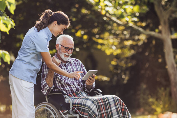 Hombre mayor en silla de ruedas mostrando algo en la tableta a la enfermera joven cuidador detrás de él en el parque - Foto, imagen