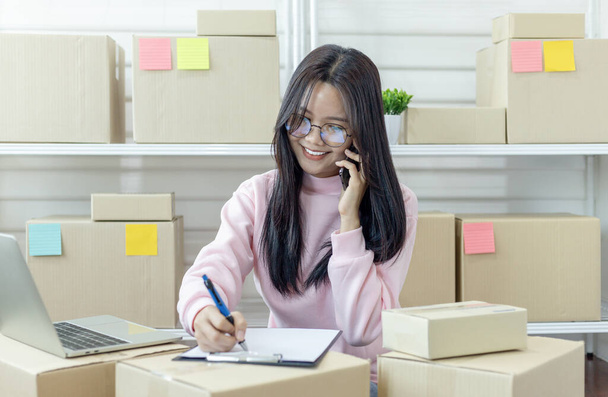 Fiatal ázsiai nő beszél okostelefonon, és ellenőrzi a termék vásárlási megrendelés és előkészíti a csomagot a szállítás az ügyfelek számára. Termékek online értékesítése. Online vásárlás. Online üzlet, Munka otthon koncepció. - Fotó, kép