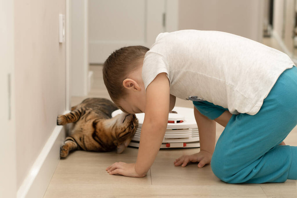Маленький хлопчик, 4 роки, і красива, червона, домашня, леопардова кішка бенгальської породи розважаються, граючи в домашньому інтер'єрі з роботом-пилососом, який прибирає квартиру. - Фото, зображення