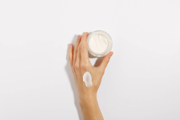 kobieca delikatna ręka z rozmazanym śmietaną szklanym słoikiem kremu do twarzy na białym, izolowanym tle. pojęcie produktów kosmetycznych, pielęgnacji skóry, kosmetologii - Zdjęcie, obraz