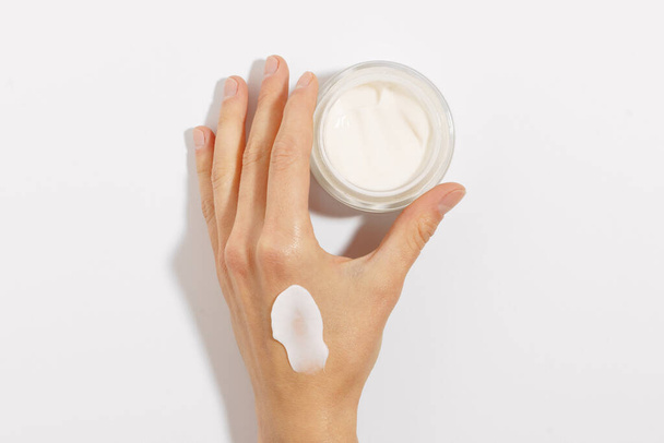 ręka żeńska z rozmazem białego kremu trzymającego szklany słoik kremu nawilżającego na białym, izolowanym tle. pojęcie produktów kosmetycznych. makieta dla Twojego projektu - Zdjęcie, obraz