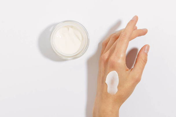 Fragile female hand with smear of cream and glass jar of facial moisturizer on white isolated background. Concept of skin care cosmetics, beauty products and cream sample - Photo, Image