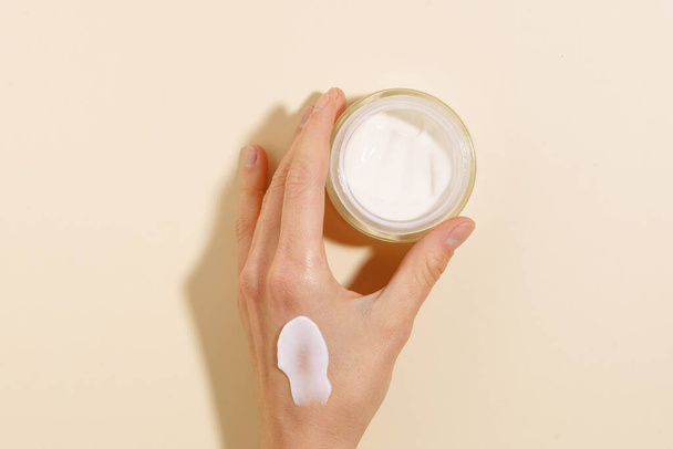 Vrouwelijke hand met een uitstrijkje crème met een glazen pot moisturizer op een beige geïsoleerde achtergrond. Concept van huidverzorging, schoonheidsproducten. Afbeelding voor uw ontwerp - Foto, afbeelding